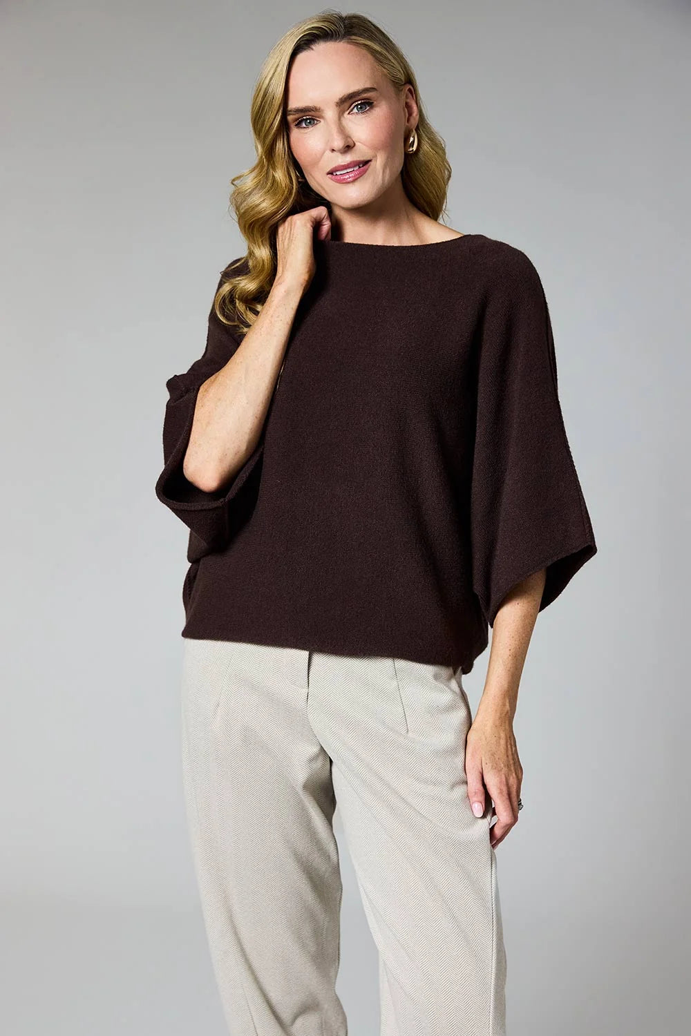 A woman modeling a dark brown boat-neck knit top with wide three-quarter sleeves, paired with light beige trousers, against a solid gray studio background by Weirs Gifts