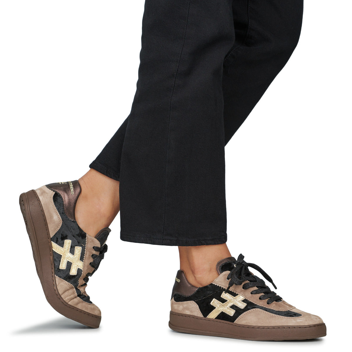 A person wearing black trousers and brown and beige sneakers with black suede panels and a gold cross logo, shown from the calves down against a white background at Weirs Gifts