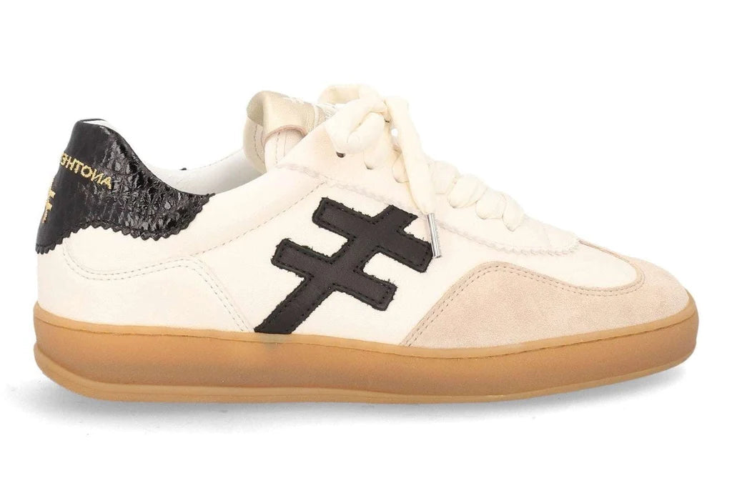 A low-top sneaker featuring an off-white leather and beige suede upper, a prominent black cross logo on the side, a black textured heel tab with gold lettering, and a thick gum sole at Weirs Gifts