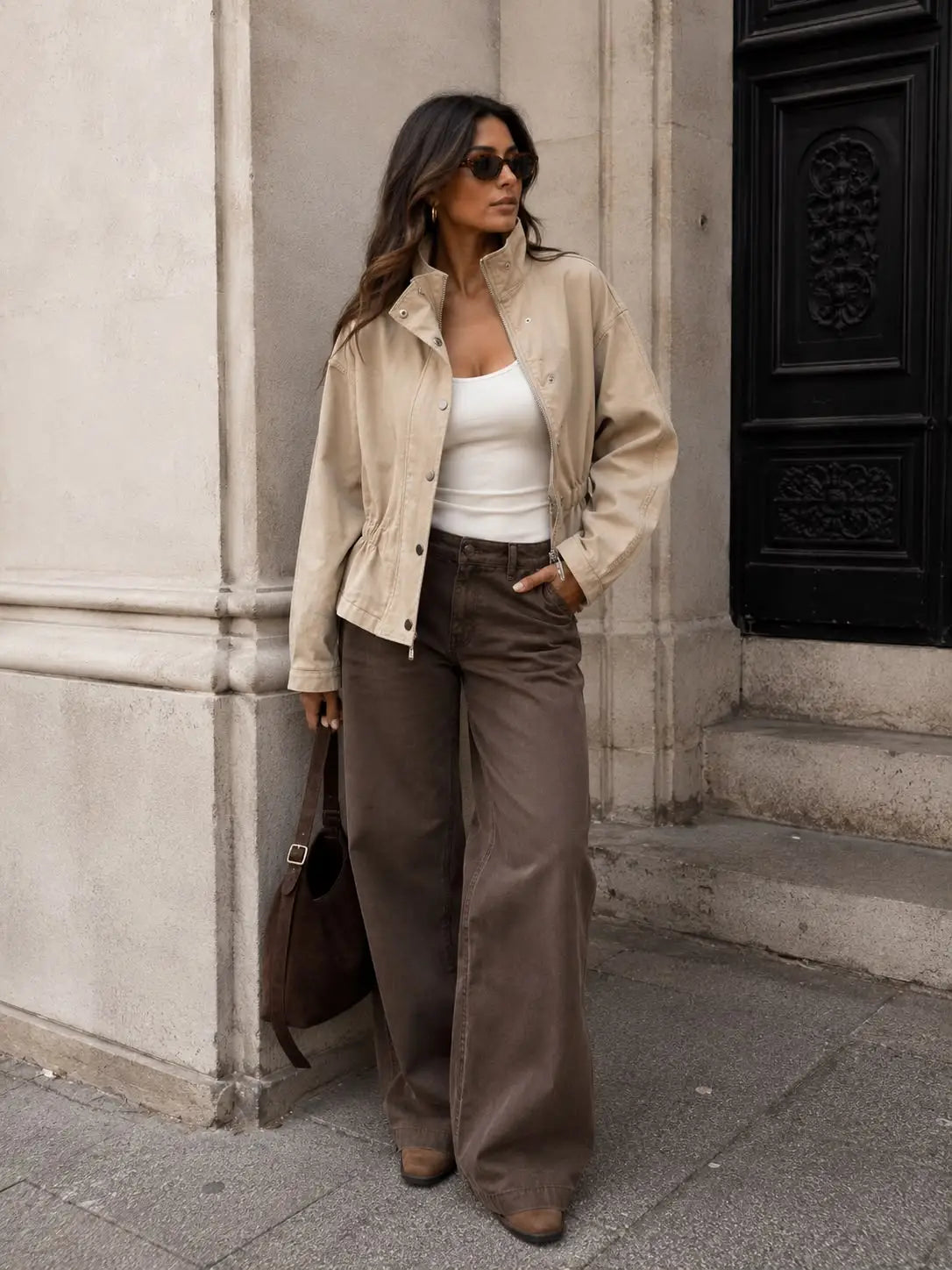 A woman stands on a city sidewalk wearing a beige utility jacket over a white tank top, brown wide-leg pants, and sunglasses, holding a brown handbag by Weirs Gifts