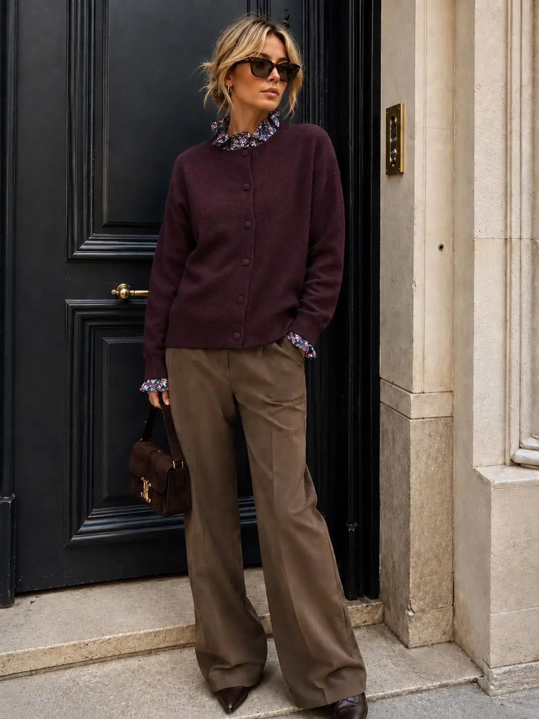 A woman stands in front of a black door wearing a dark burgundy button-up cardigan over a floral collared shirt, olive brown wide-leg trousers, and dark pointed shoes, holding a brown handbag by Weirs Gifts