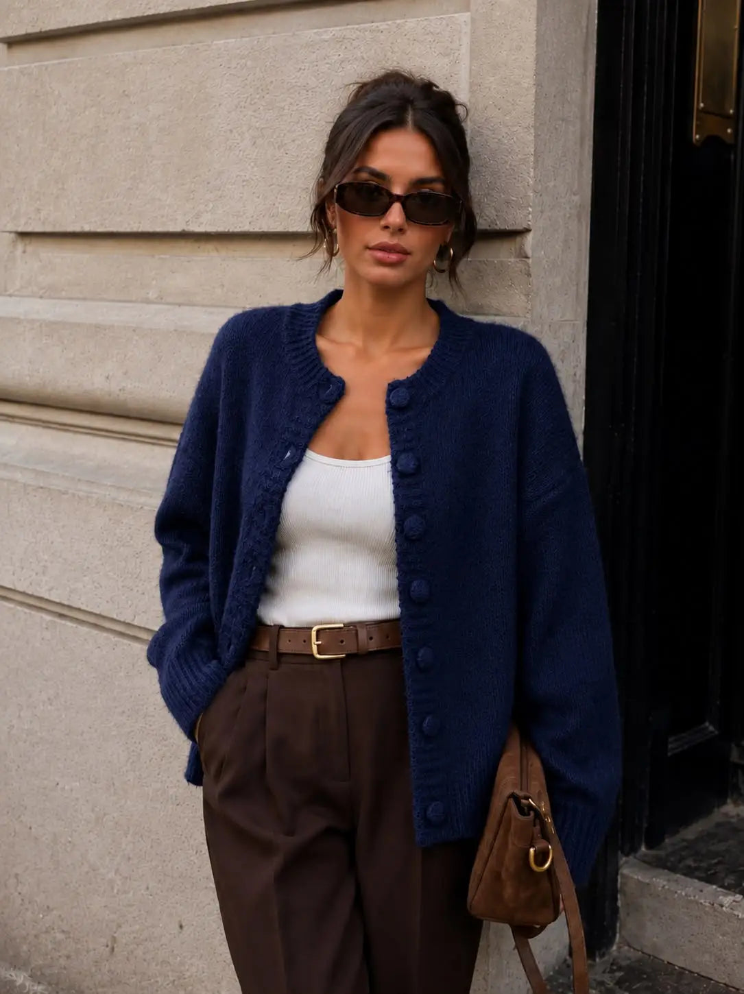 A woman wearing a navy blue button-front cardigan over a white tank top, paired with brown trousers and holding a brown shoulder bag, standing against a beige stone wall by Weirs Gifts