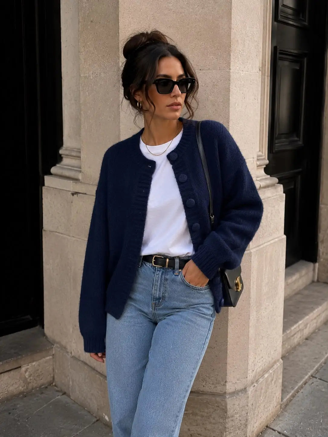 A woman wearing a navy blue knit cardigan over a white t-shirt, light-wash blue jeans, and black sunglasses, standing outdoors against a beige stone building by Weirs Gifts