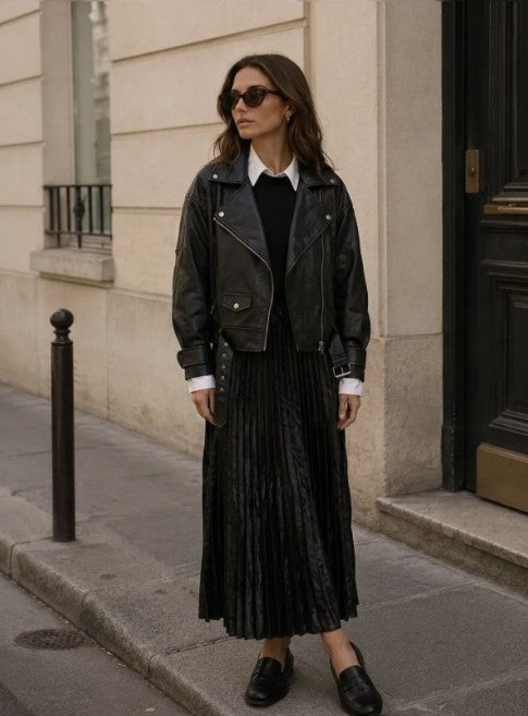 A woman stands on a city sidewalk wearing a black leather moto jacket over a black top and a long black pleated skirt, with black loafers and sunglasses at Weirs Gifts