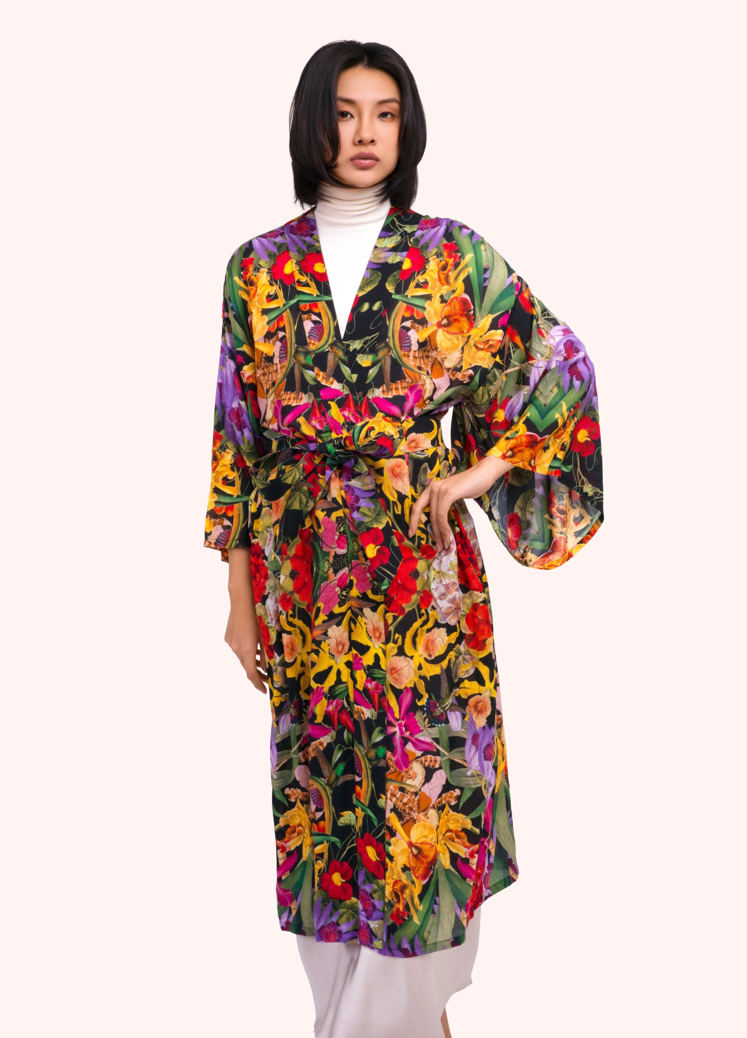 A model wearing a vibrant, multicolored floral kimono robe over a white turtleneck and white trousers, set against a solid pale pink background by Weirs Gifts