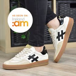 A person wearing cream-colored sneakers with black side stripes, black heel tabs, white laces, and gum soles, standing on a wooden floor. A circular badge in the top left says 'AS SEEN ON Ireland 2am' at Weirs Gifts