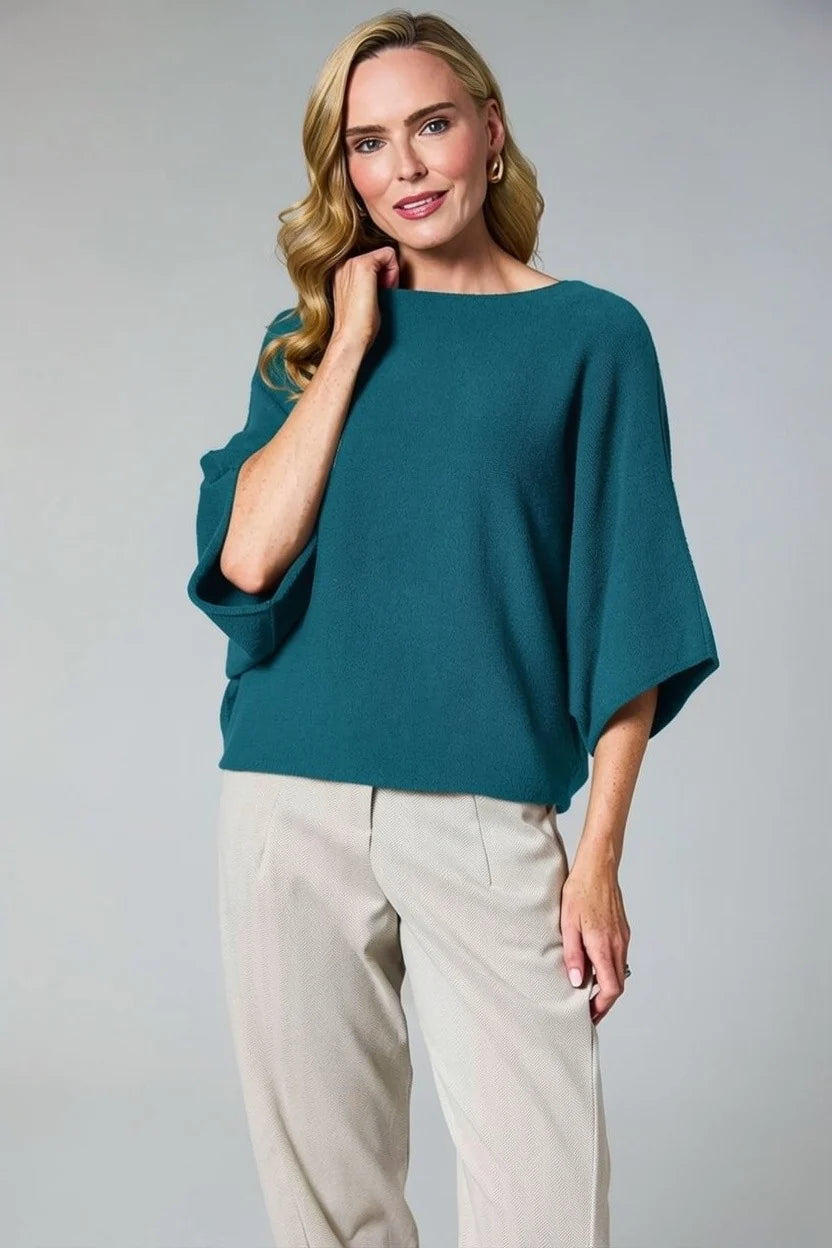A woman modeling a teal wide-sleeve boatneck top with beige trousers, standing against a neutral gray studio background by Weirs Gifts