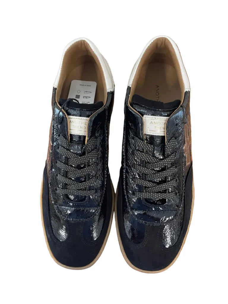A black leather low-top sneaker with a glossy patent finish, featuring a metallic bronze geometric logo on the side, a cream-colored heel tab with gold lettering, and a gum brown sole at Weirs Gifts