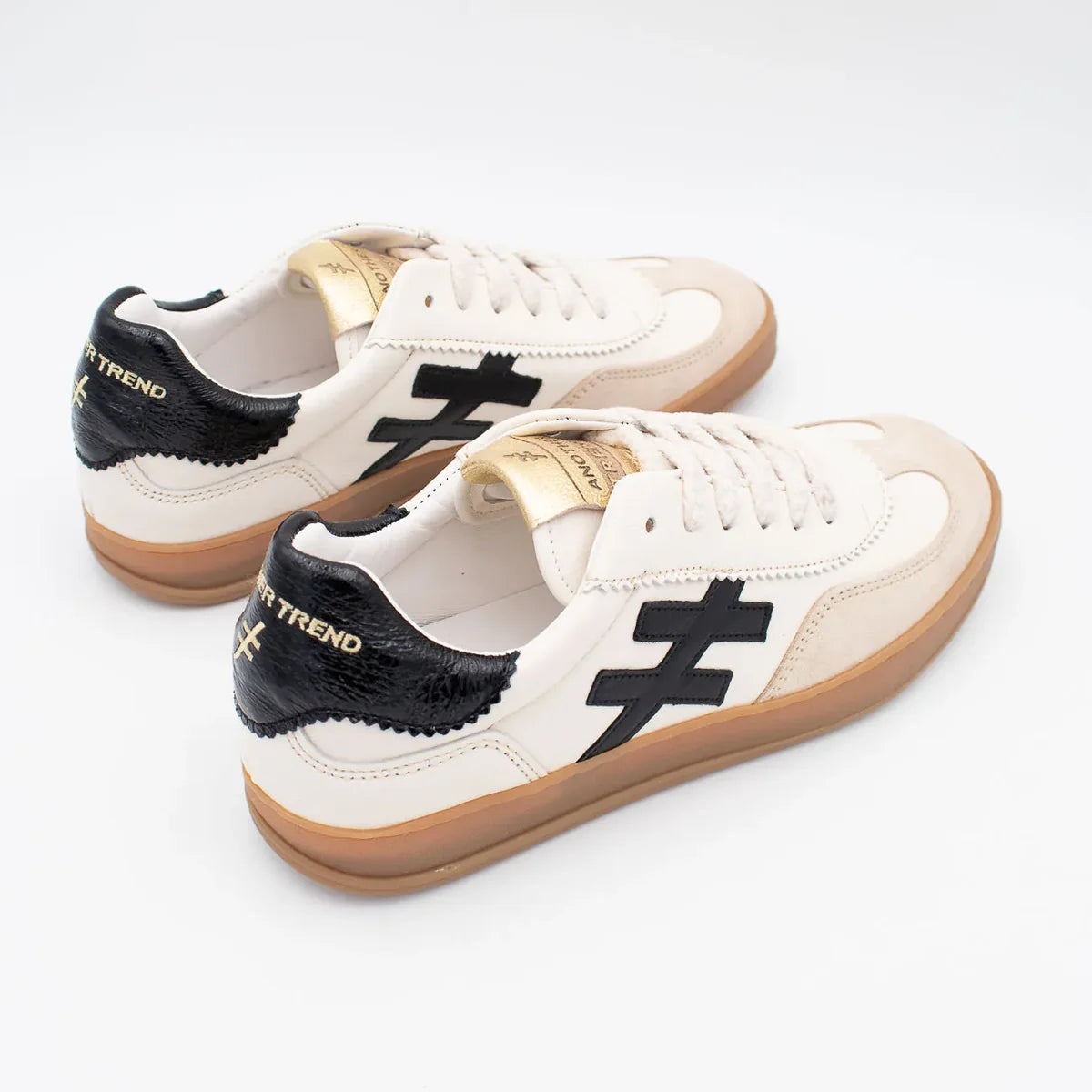 A low-top sneaker featuring an off-white leather and beige suede upper, a prominent black cross logo on the side, a black textured heel tab with gold lettering, and a thick gum sole at Weirs Gifts