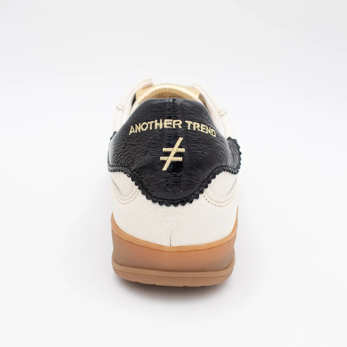 A low-top sneaker featuring an off-white leather and beige suede upper, a prominent black cross logo on the side, a black textured heel tab with gold lettering, and a thick gum sole at Weirs Gifts
