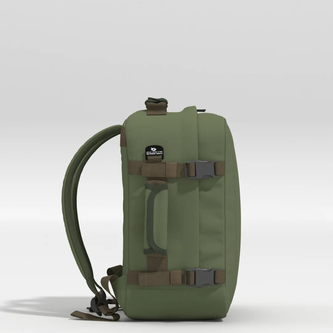 An olive green tactical backpack shown from the side against a light gray studio background. The backpack features brown webbing straps with gray plastic buckles, a clear front window, and a black brand patch.