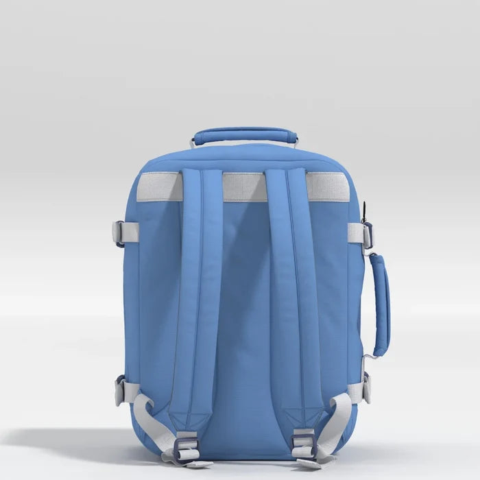 A light blue backpack shown from the back, featuring padded shoulder straps, adjustable gray buckles, reflective gray panels, and a top handle, set against a neutral light gray background.