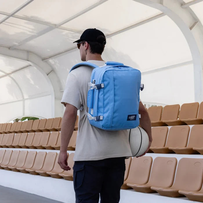 A man wearing a black baseball cap and a light beige t-shirt walks through an indoor stadium with rows of beige seats, carrying a white basketball in one hand and wearing a bright blue backpack on his back.