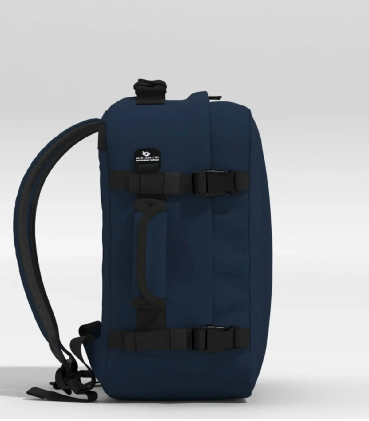 A dark navy blue backpack shown from the side against a light gray studio background. The backpack features black straps, buckles, and a black top handle, with a small white logo patch on the upper front.