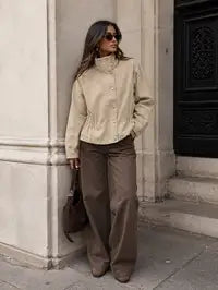 A woman stands outdoors against a stone building corner wearing a beige cropped jacket with a stand collar and front buttons, brown wide-leg pants, sunglasses, and carrying a brown shoulder bag.