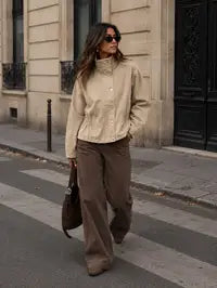 A woman walking on a city street wearing a beige mock-neck button-up shirt, brown wide-leg trousers, and carrying a dark brown tote bag.