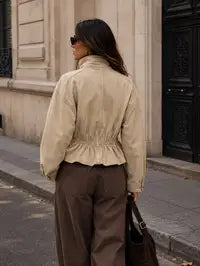 A woman photographed from behind wearing a beige cropped jacket with a gathered waist and brown wide-leg trousers, standing on a city sidewalk in front of a building with a dark door by Weirs Gifts