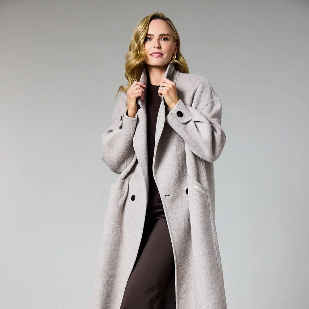 A woman modeling a long, light gray double-breasted coat over a dark brown top and wide-leg trousers, standing against a neutral gray studio background at Weirs Gifts