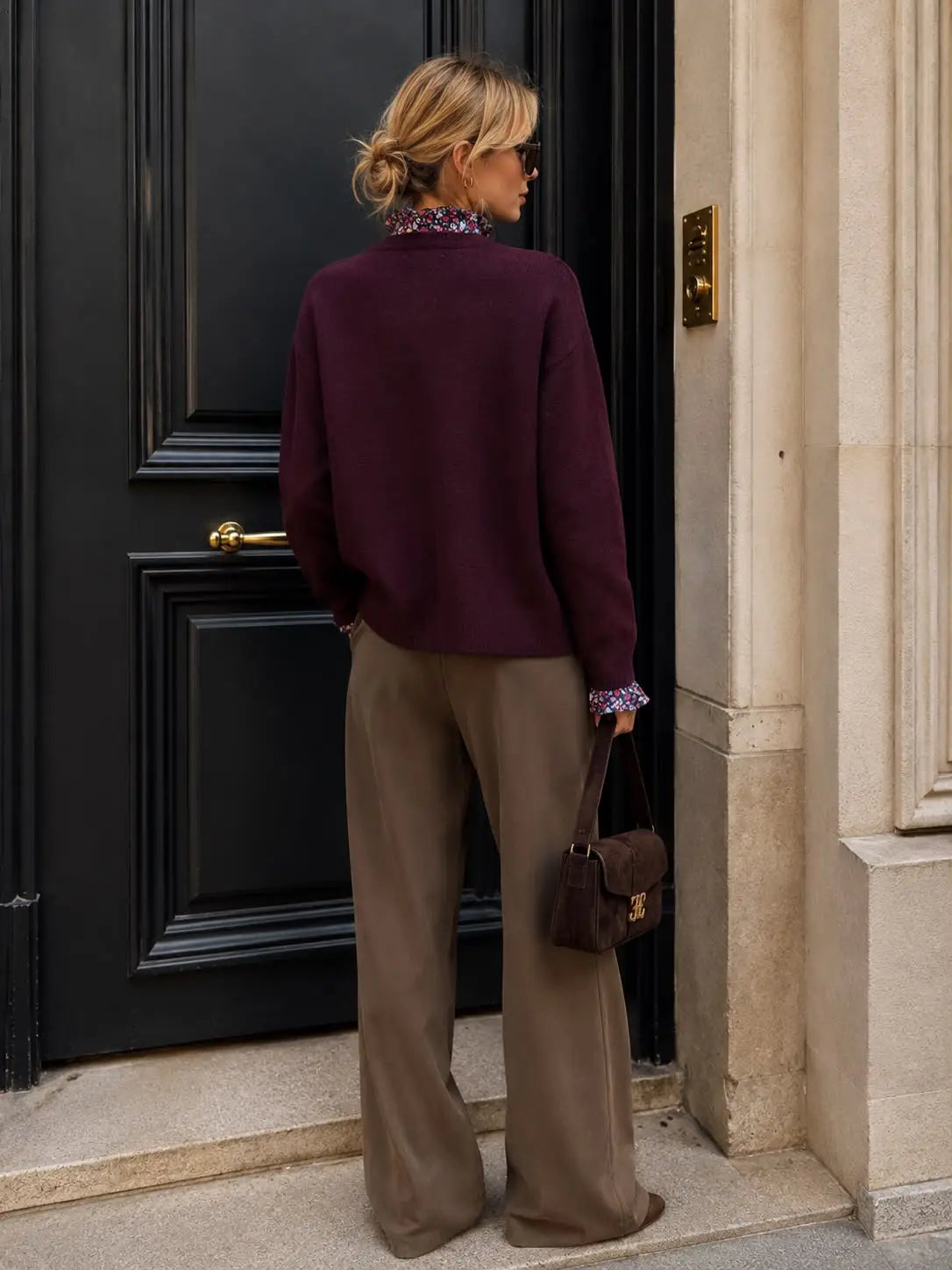 A woman stands in front of a black door wearing a deep burgundy knit sweater, wide-leg taupe trousers, and holding a brown handbag.