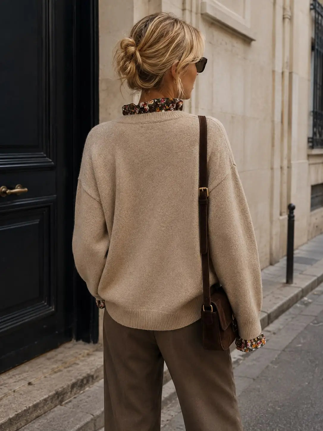 A woman photographed from behind on a city sidewalk, wearing a beige knit sweater with floral trim at the collar and cuffs, brown trousers, and carrying a brown shoulder bag by Weirs Gifts