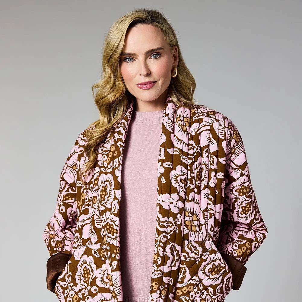 A woman modeling a brown and pink floral patterned jacket over a light pink sweater and blue jeans, photographed against a neutral gray studio background at Weirs Gifts