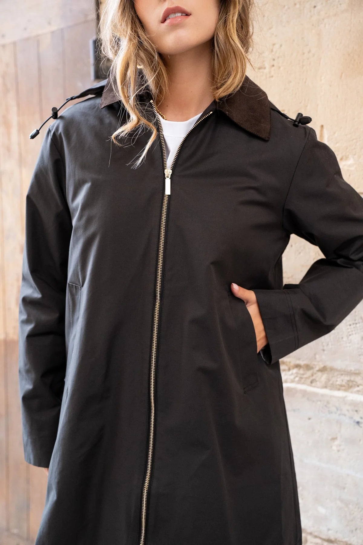 A woman wearing a black trench coat with a brown collar, standing against a textured stone wall at Weirs Gifts