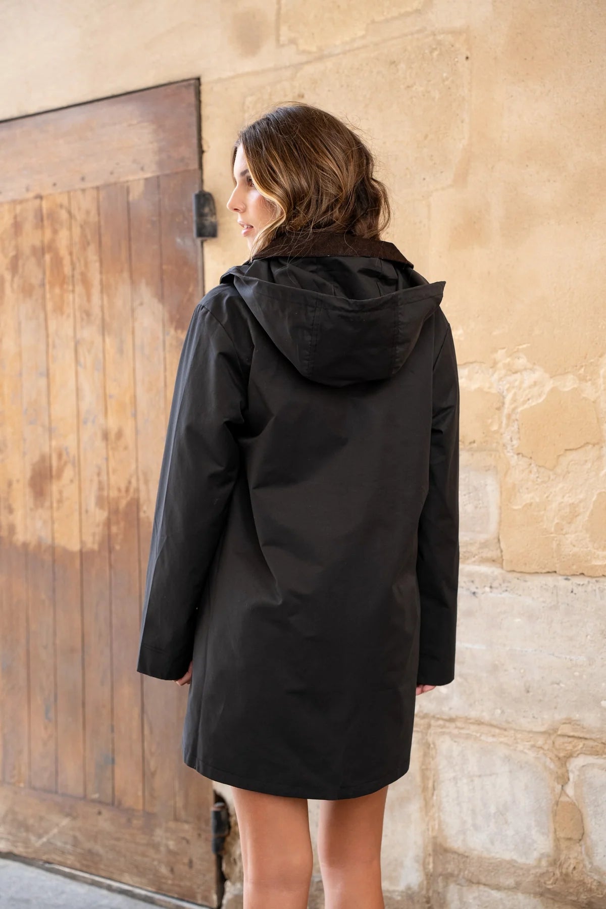 A woman wearing a black hooded coat, photographed from behind against a beige stone wall and wooden door at Weirs Gifts