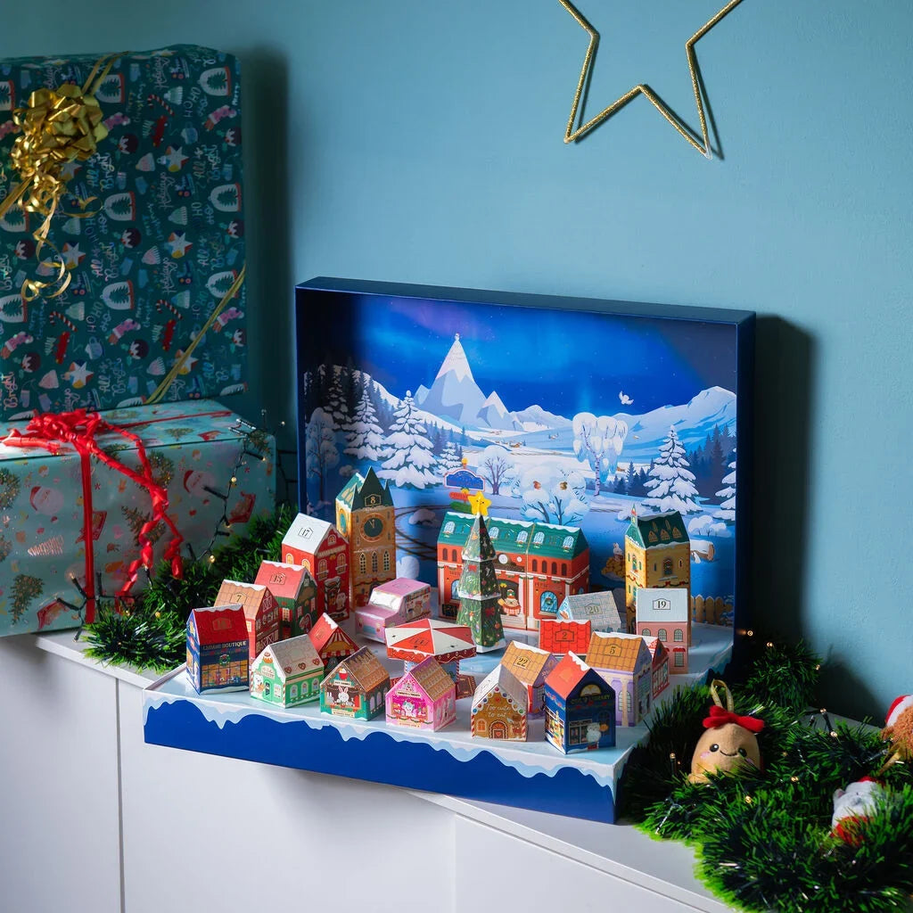 A decorative advent calendar set shaped like a snowy village, displayed on a shelf in a holiday setting with wrapped gifts and greenery against a blue wall at Weirs Gifts.