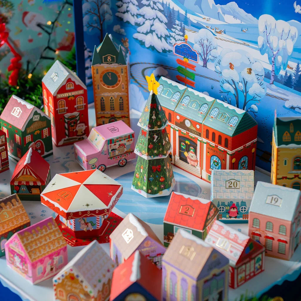 A festive Christmas advent-style scene with colorful paper house models, a decorated Christmas tree, a pink toy car, and a winter village backdrop featuring snow-covered trees and buildings at Weirs Gifts.