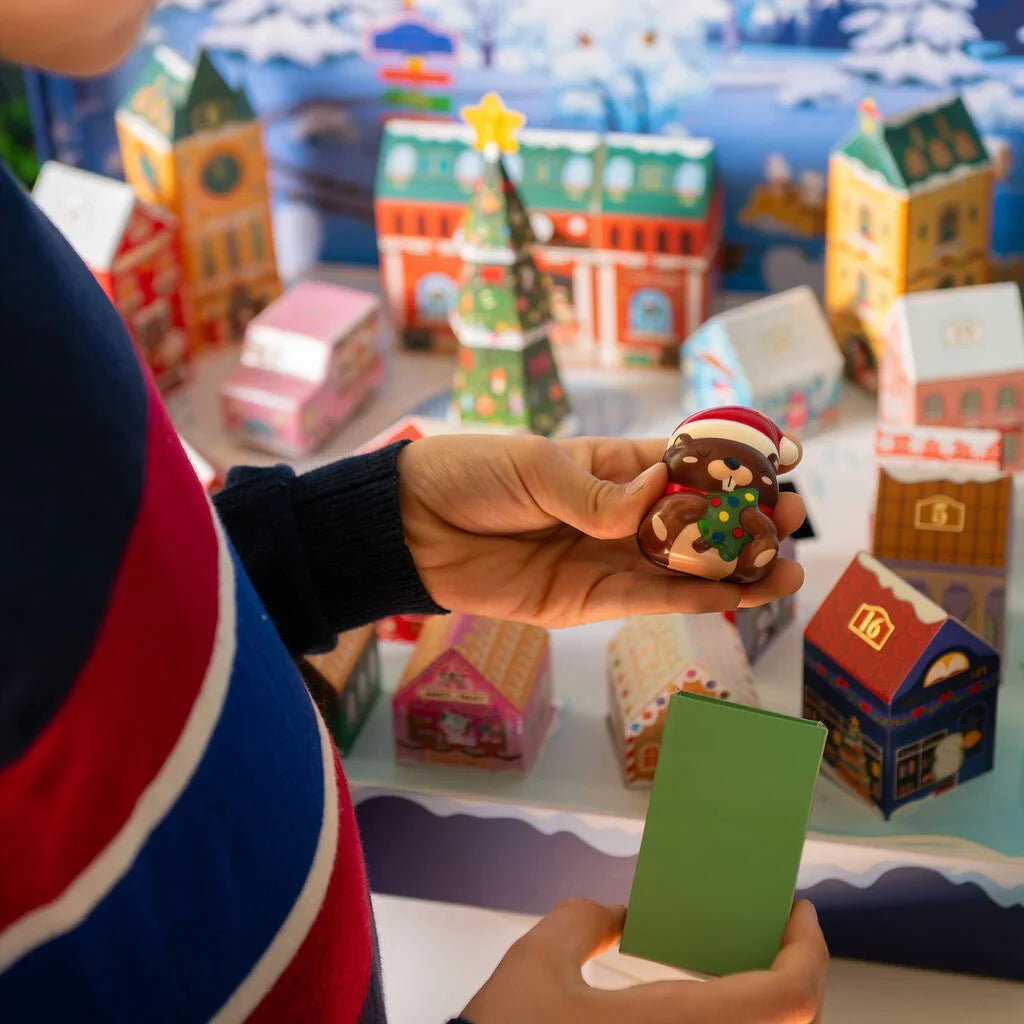 A person holds a small Christmas-themed brown bear figurine wearing a Santa hat in front of a colorful miniature village display of houses and trees at Weirs Gifts