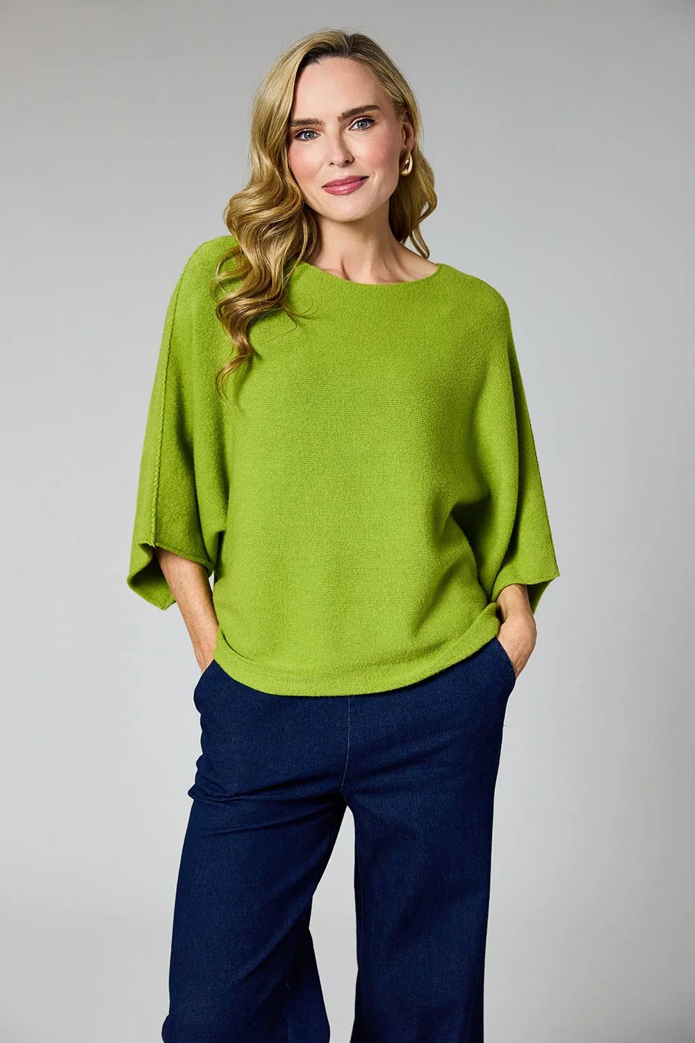 A woman modeling a bright lime green knit top with wide three-quarter sleeves and dark navy wide-leg pants, standing against a neutral gray studio background at Weirs Gifts