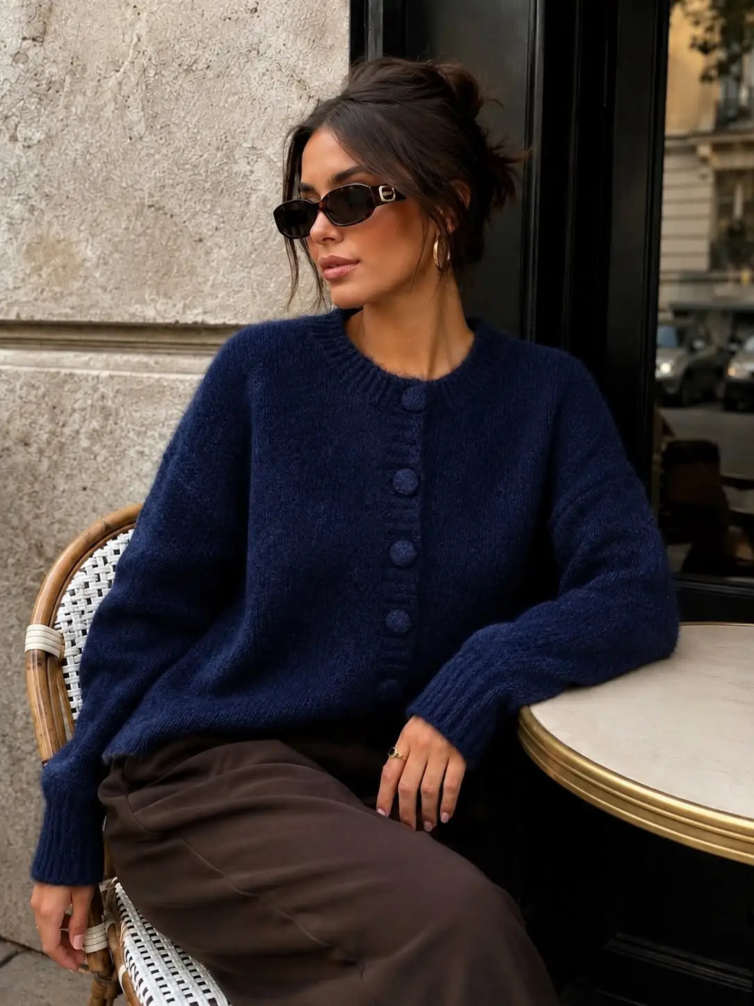 A woman wearing a dark navy blue fuzzy knit cardigan sweater with large buttons, styled with brown trousers, sitting on a cafe chair next to a table.