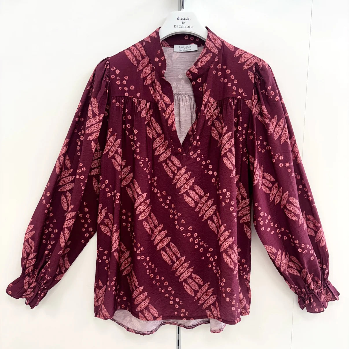 A long-sleeve blouse in deep burgundy with a pinkish-red leaf and dot pattern, hanging on a white hanger against a white wall by Weirs Gifts