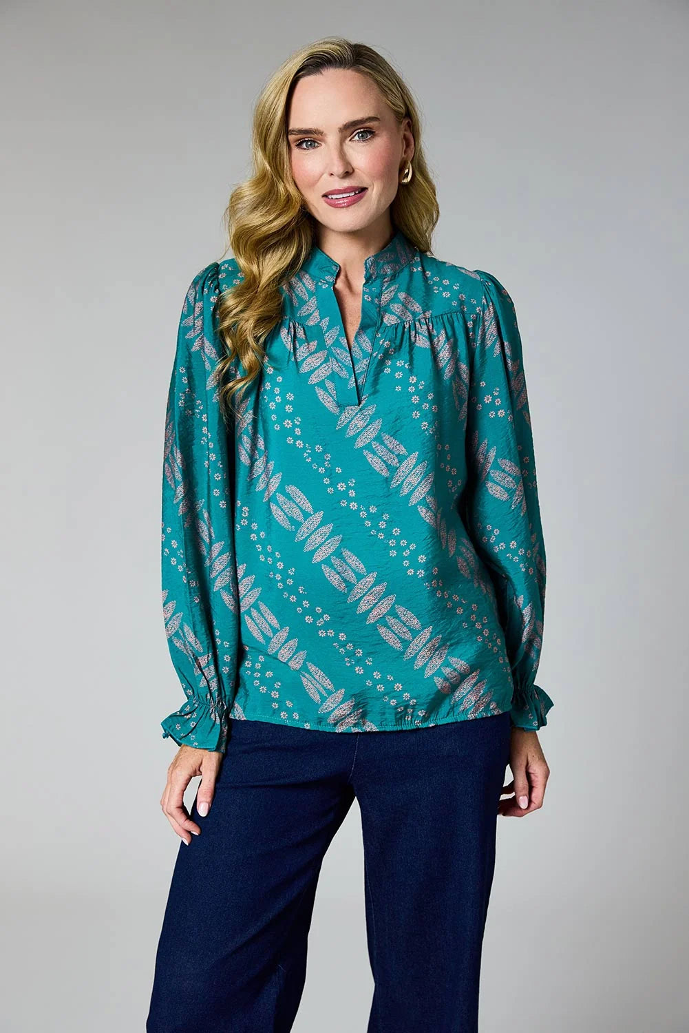 A woman modeling a teal long-sleeve blouse with a gray geometric pattern, paired with dark navy pants, against a neutral gray studio background by Weirs Gifts