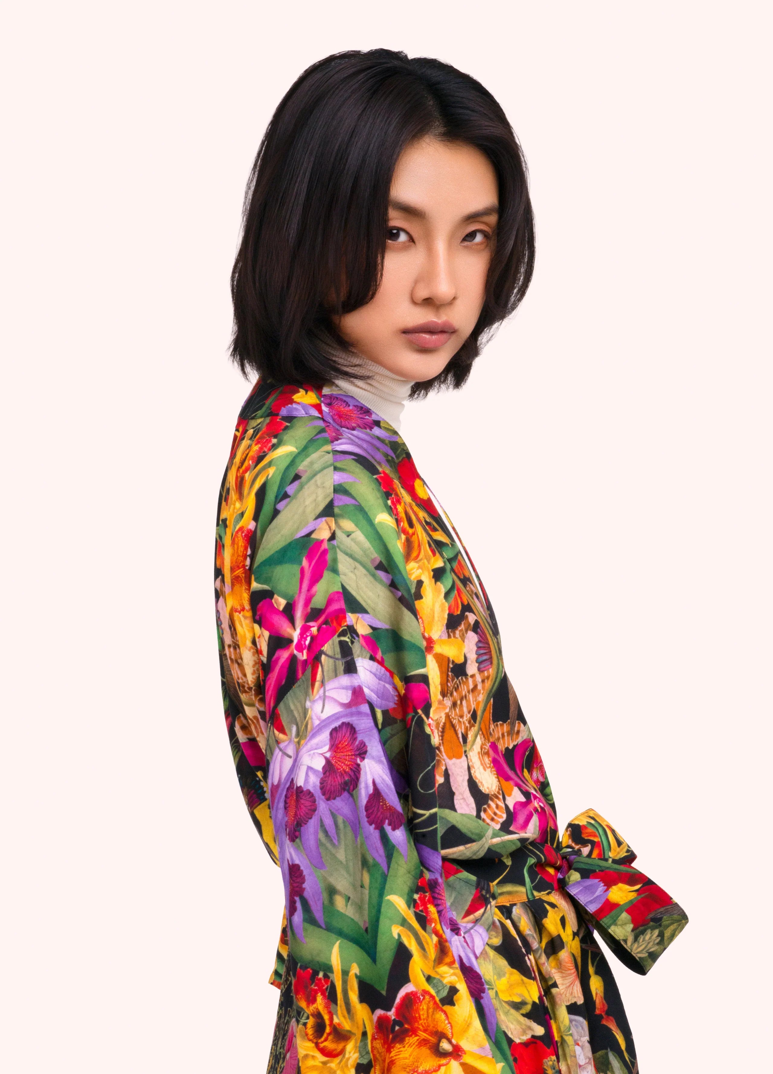 A model with short dark hair is shown from a side-back angle wearing a vibrant, multicolored floral print kimono-style robe with a tie belt, set against a solid pale pink background by Weirs Gifts