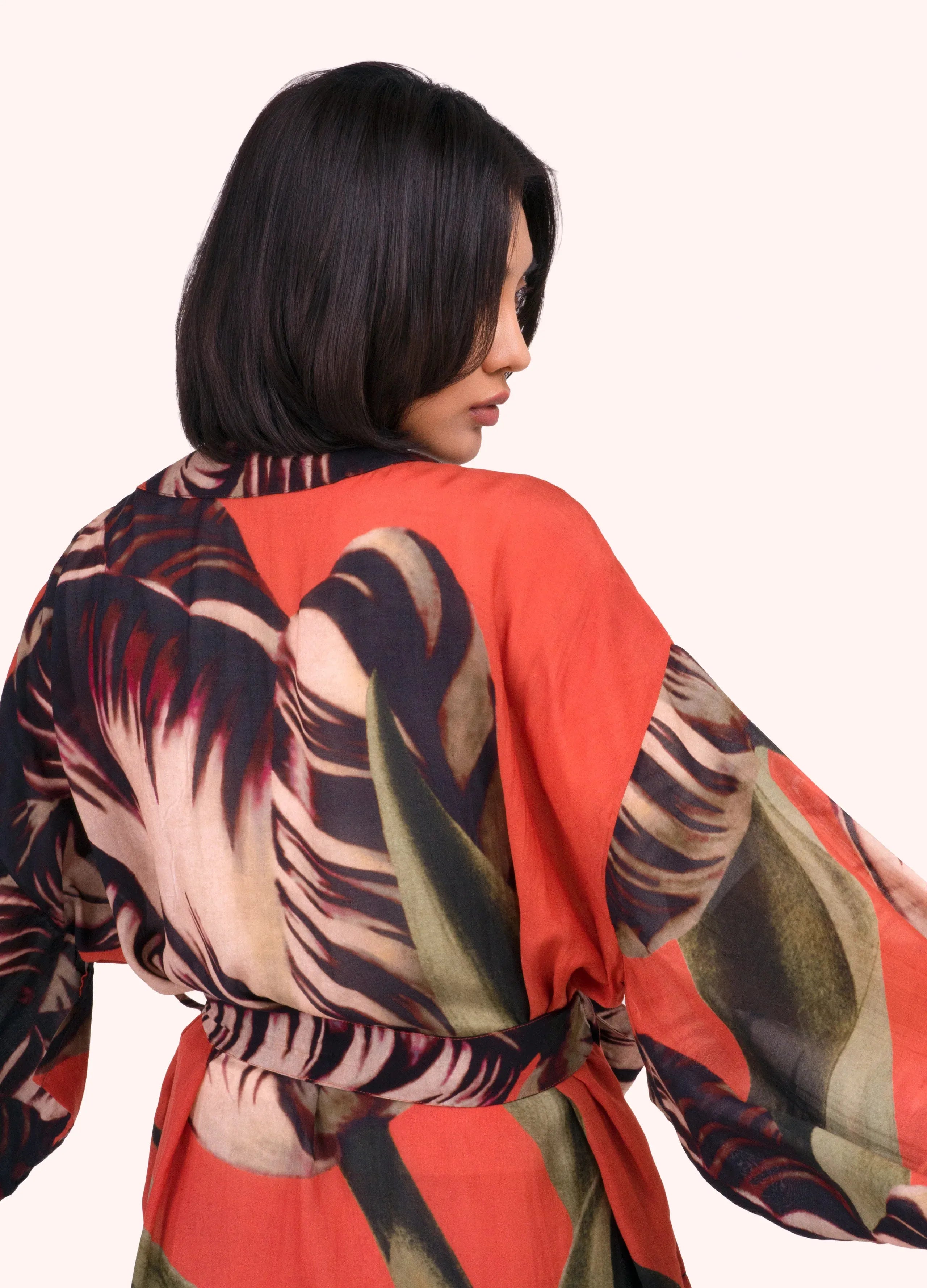 A woman shown from the back wearing a lightweight wrap or kimono-style top with a bold tropical leaf print in coral red, black, beige, and olive green on a plain light background by Weirs Gifts