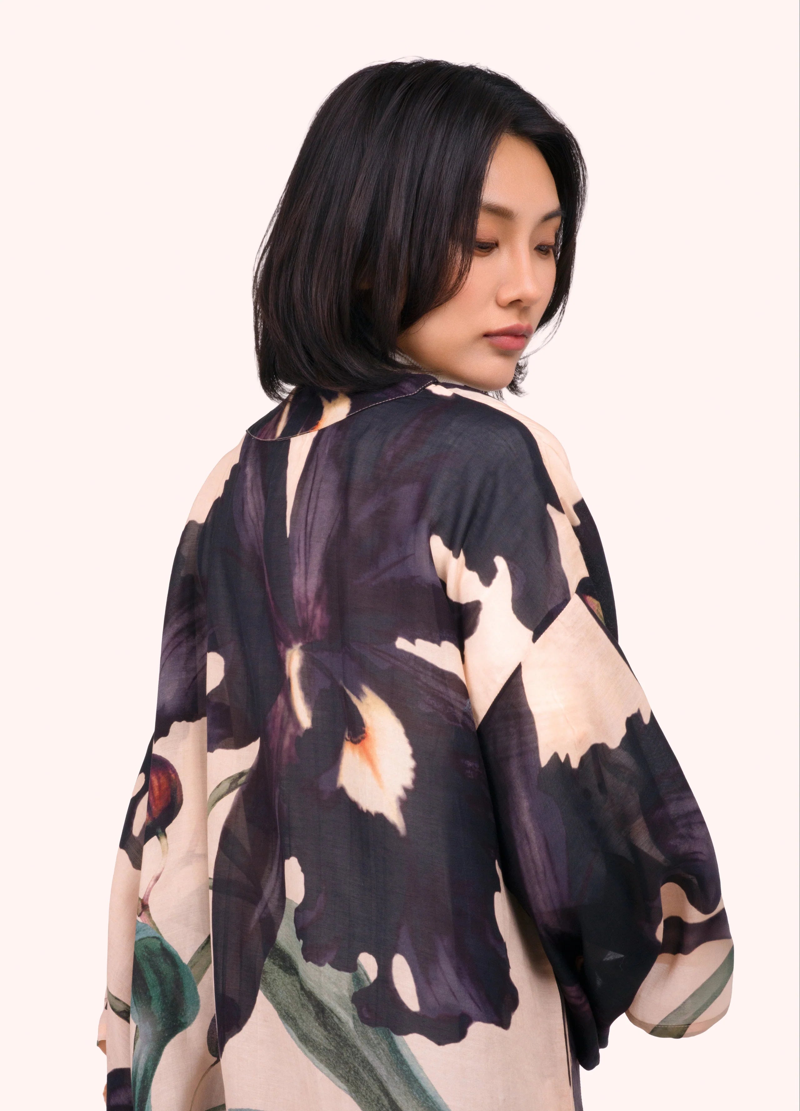 A woman with a short dark bob is shown from the back wearing a loose, wide-sleeved kimono-style top with a large floral print in dark purple, beige, and green on a light cream background by Weirs Gifts