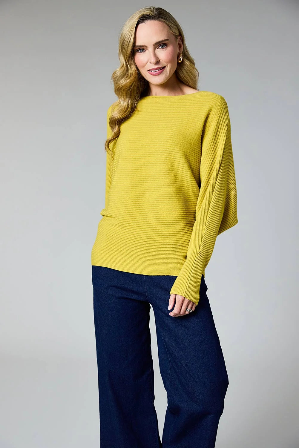 A woman modeling a bright yellow ribbed knit sweater with wide boat neckline and dolman sleeves, paired with dark navy wide-leg pants, against a neutral gray studio background by Weirs Gifts