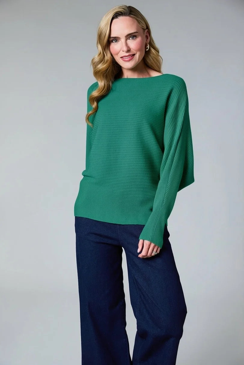 A woman modeling a green ribbed knit sweater with long draped sleeves, paired with dark navy wide-leg pants, against a neutral gray studio background.