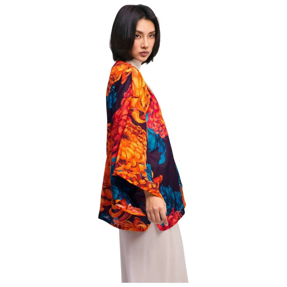 A model wearing a vibrant, multicolored floral kimono-style top with orange, red, blue, and black patterns, paired with light beige trousers, shown from a side-back angle against a plain white background by Weirs Gifts