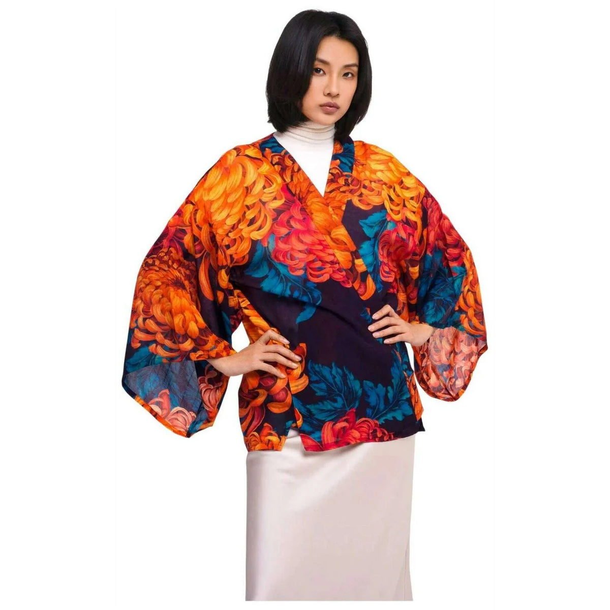 A model wearing a vibrant, multicolored kimono-style wrap top with wide sleeves, featuring a bold floral pattern in orange, red, blue, and black, styled over a white turtleneck and cream skirt against a plain white background by Weirs Gifts