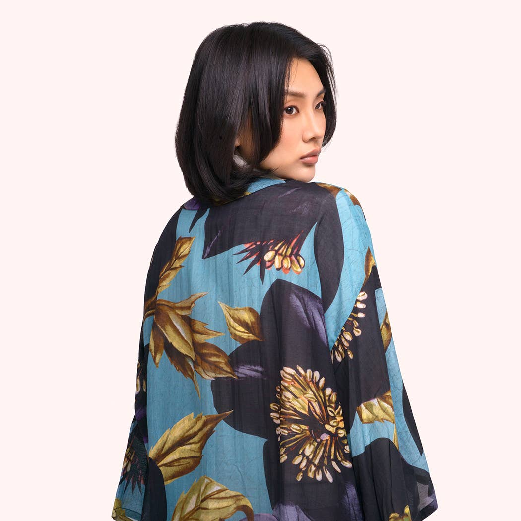 A woman with short black hair is shown from the back wearing a lightweight, flowing kimono-style top with a blue, black, and gold floral pattern, set against a solid pale pink background by Weirs Gifts