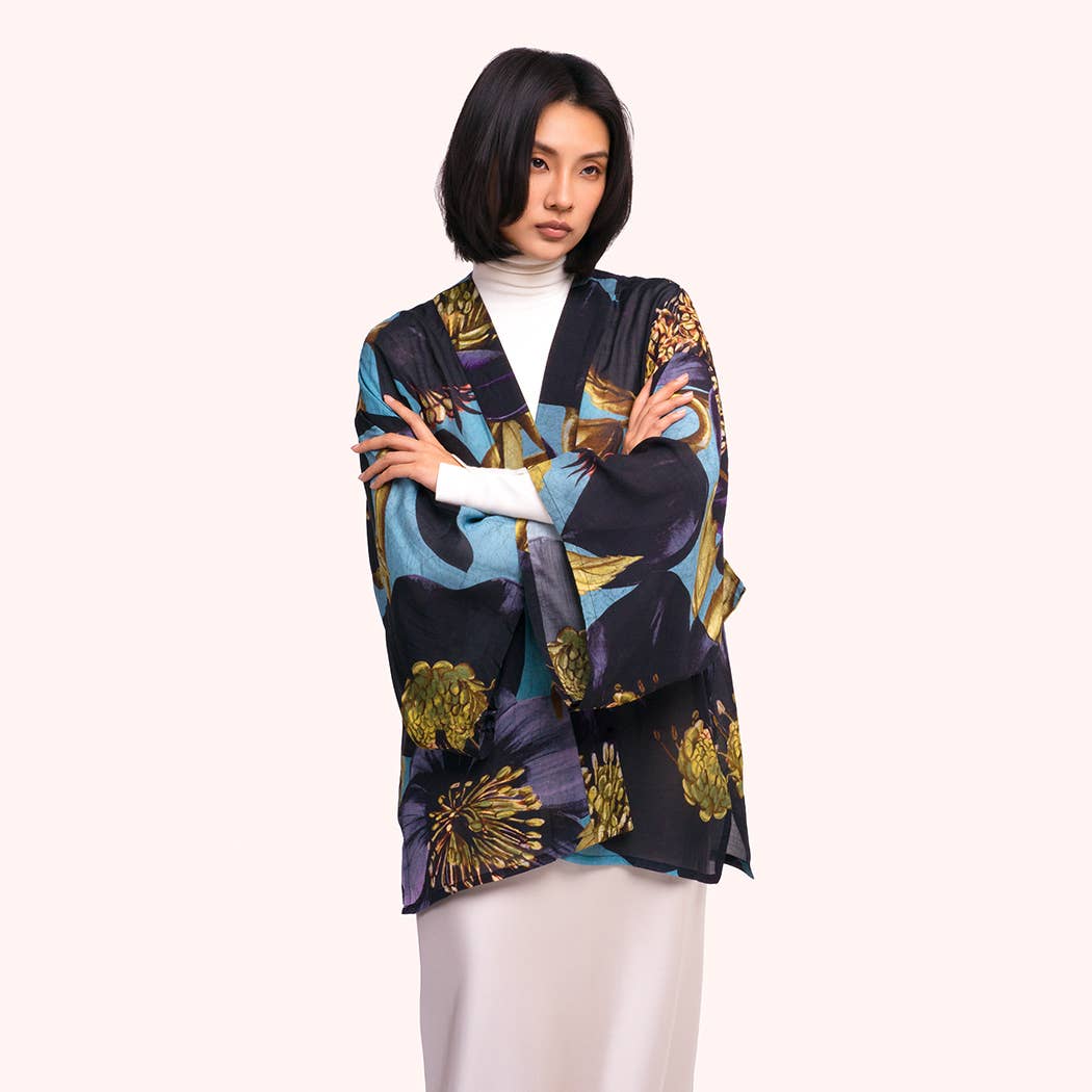 A model with short dark hair is wearing a lightweight open-front cardigan featuring a bold abstract floral pattern in navy, teal, purple, and gold. She is styled with a white mock-neck top and light beige trousers, set against a solid pale pink background by Weirs Gifts